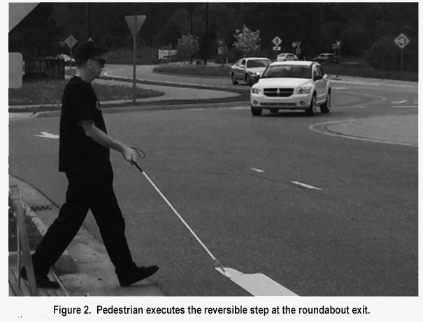 Figure 2.  Pedestrian executes the reversible step at the roundabout exit.  Picture shows man at the crosswalk, wearing black pants, shirt and baseball cap, stepping forward to put one foot at the edge of the road, and holding a white cane on front of him.  Two cars are in the roundabout to his left, and one is turning to leave the roundabout and approach his crosswalk.
